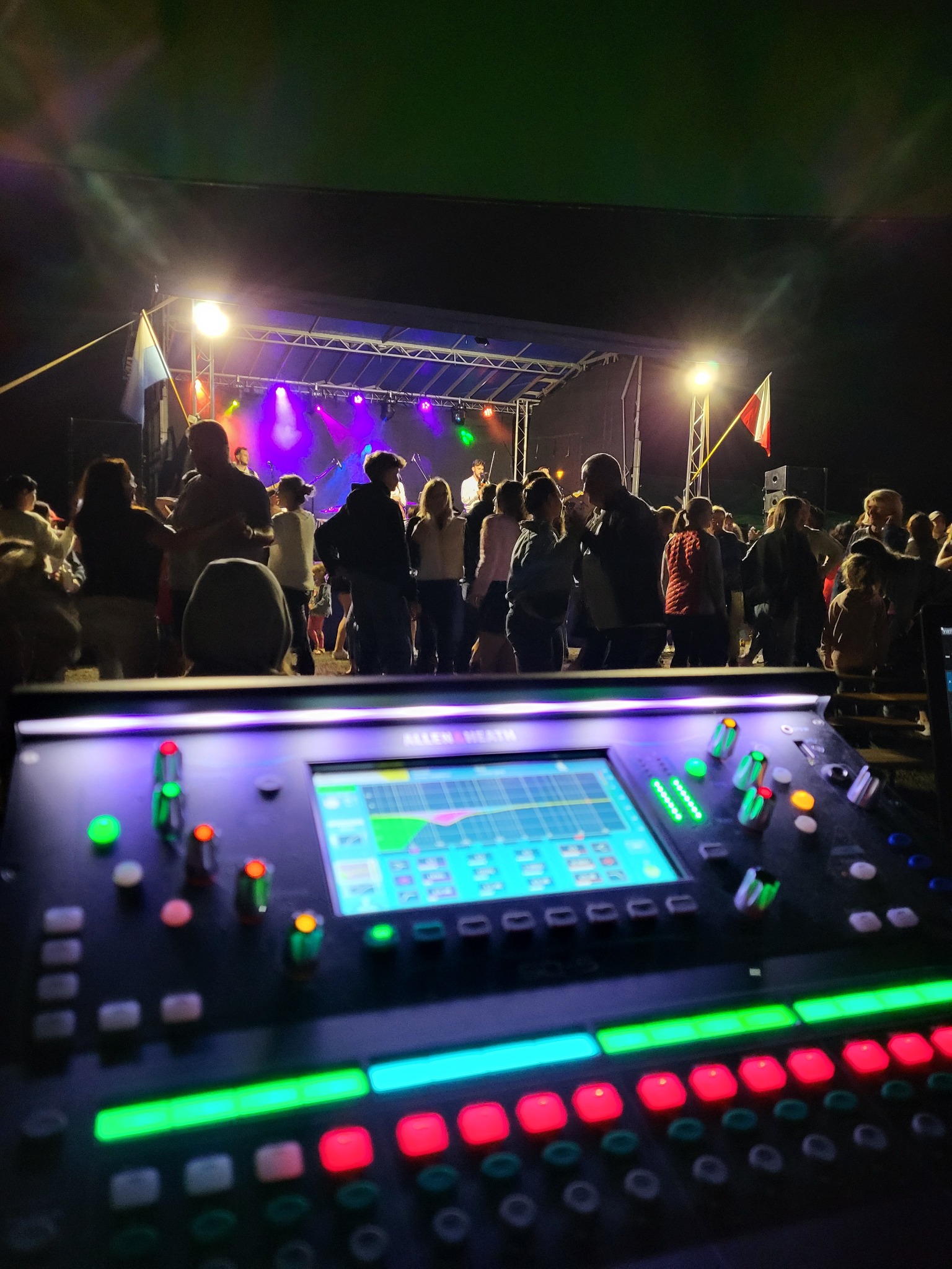Konsoleta mikserska Allen&Heath na tle sceny plenerowej z publicznością, oświetlona reflektorami. Widoczne flagi. Nocne ujęcie z bliska.