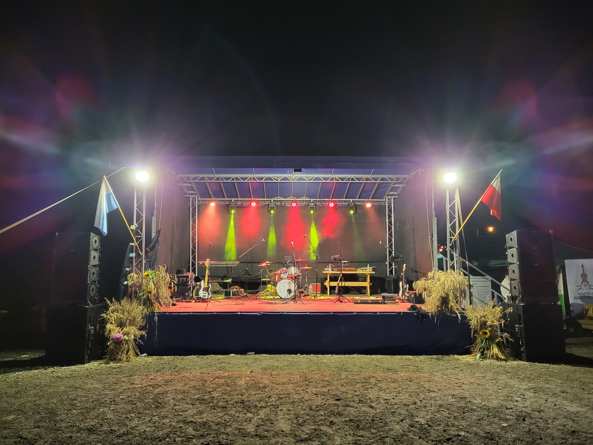 Scena plenerowa oświetlona reflektorami, z instrumentami muzycznymi i dekoracjami z kwiatów, flagi po bokach. Przygotowania do występu.