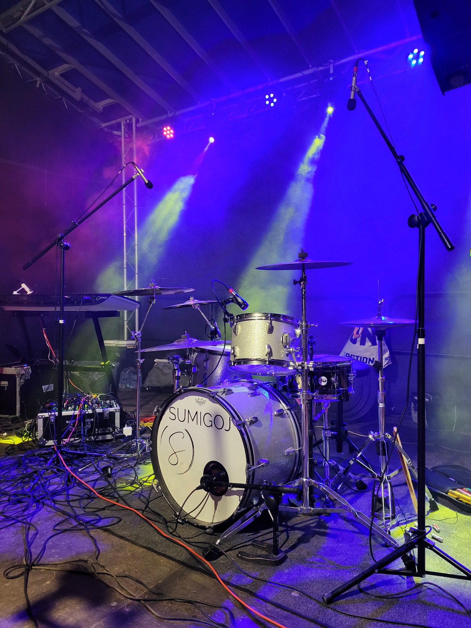 Perkusja zespołu Sumigoj na scenie w oświetleniu estradowym. Widoczne mikrofony, kable i klawisze. Dynamiczne światła w kolorach niebieskim i żółtym tworzą atmosferę koncertu.