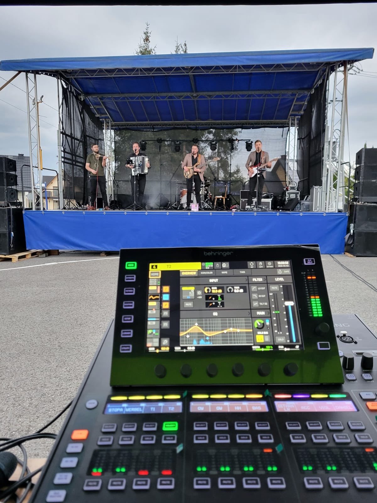 Zespół na scenie, widok zza konsolety mikserskiej Behringer. Muzycy grają na akordeonie, gitarach i skrzypcach. Scena z niebieskim tłem, nagłośnienie po bokach.