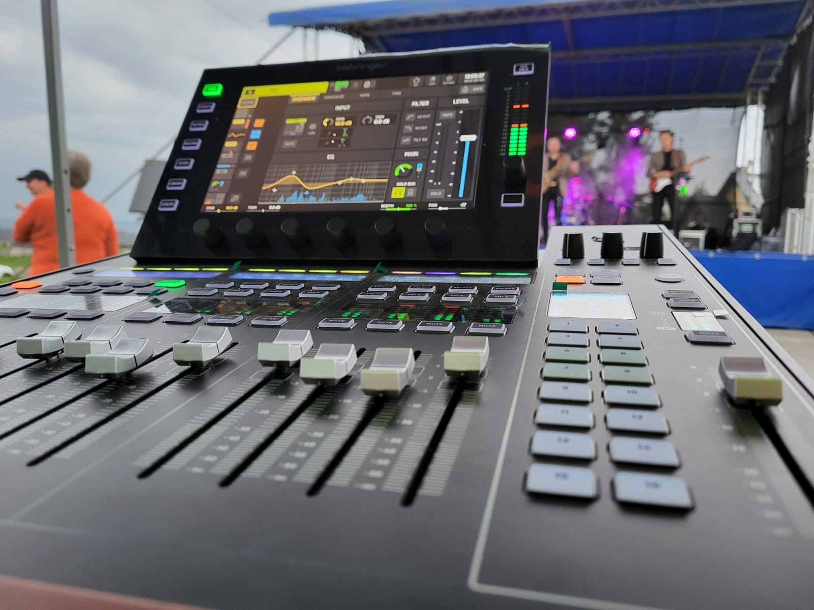 Profesjonalny mikser cyfrowy Behringer X32 z widocznym zespołem muzycznym na scenie w tle, sugerujący realizację nagłośnienia koncertowego.
