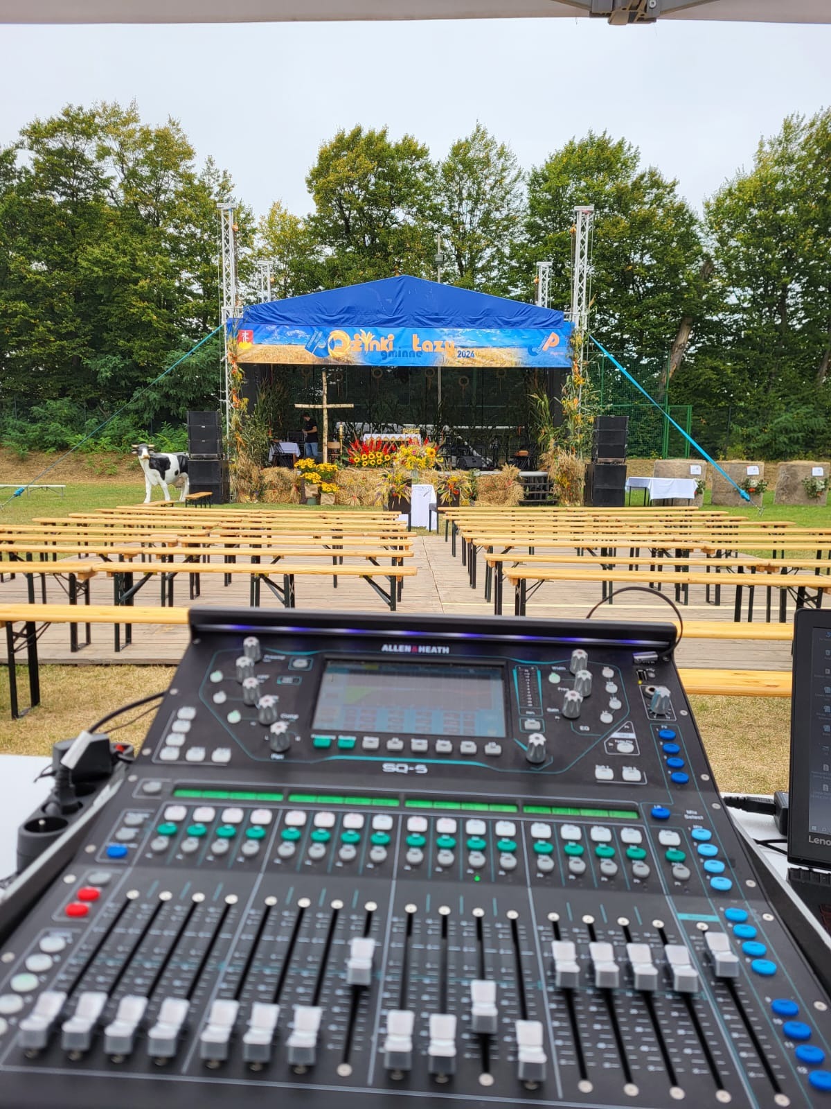 Konsoleta Allen&Heath SQ-5 na tle sceny plenerowej z dekoracjami dożynkowymi i rzędami ławek. Na lewo stoi krowa.