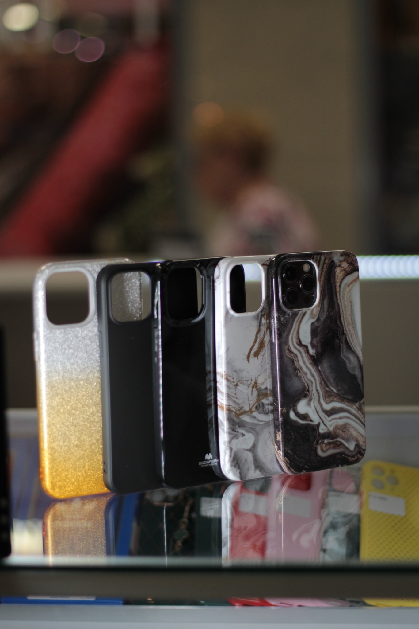 Cztery różne etui na telefon, od brokatu przechodzącego w złoto po marmurkowy wzór, prezentowane w witrynie sklepu w Białymstoku. Widoczne odbicia w szklanej powierzchni ekspozycyjnej.