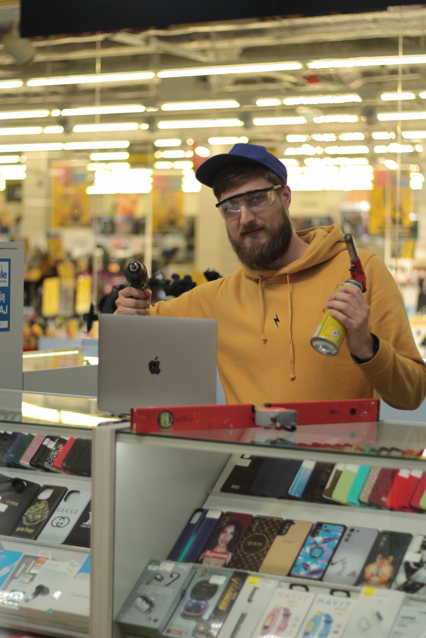 Mężczyzna w okularach ochronnych i bluzie z kapturem trzymający w jednej ręce wiertarkę, a w drugiej palnik gazowy, stojący za ladą z etui na telefony i laptopem Apple.