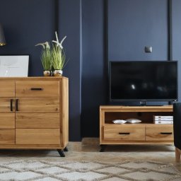 IGA Meble - Drewniana komoda z sześcioma szufladami i czarnymi uchwytami, obok szafka RTV z telewizorem na tle granatowej ściany z dekoracyjnymi listwami, dywan w geometryczne wzory.