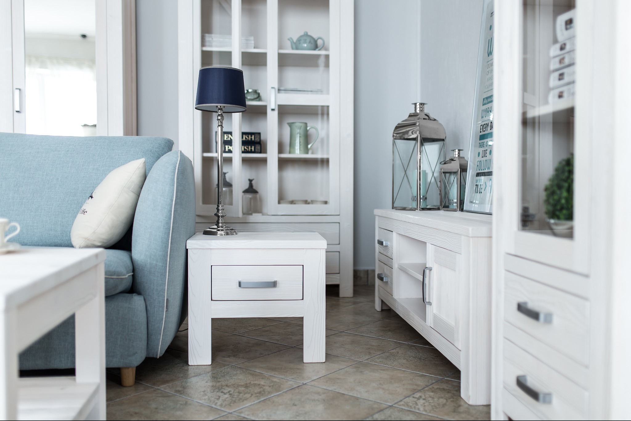 Jasne wnętrze pokoju dziennego z błękitną sofą, białymi meblami drewnianymi w stylu skandynawskim, lampą stołową z granatowym abażurem i dekoracyjnymi lampionami.