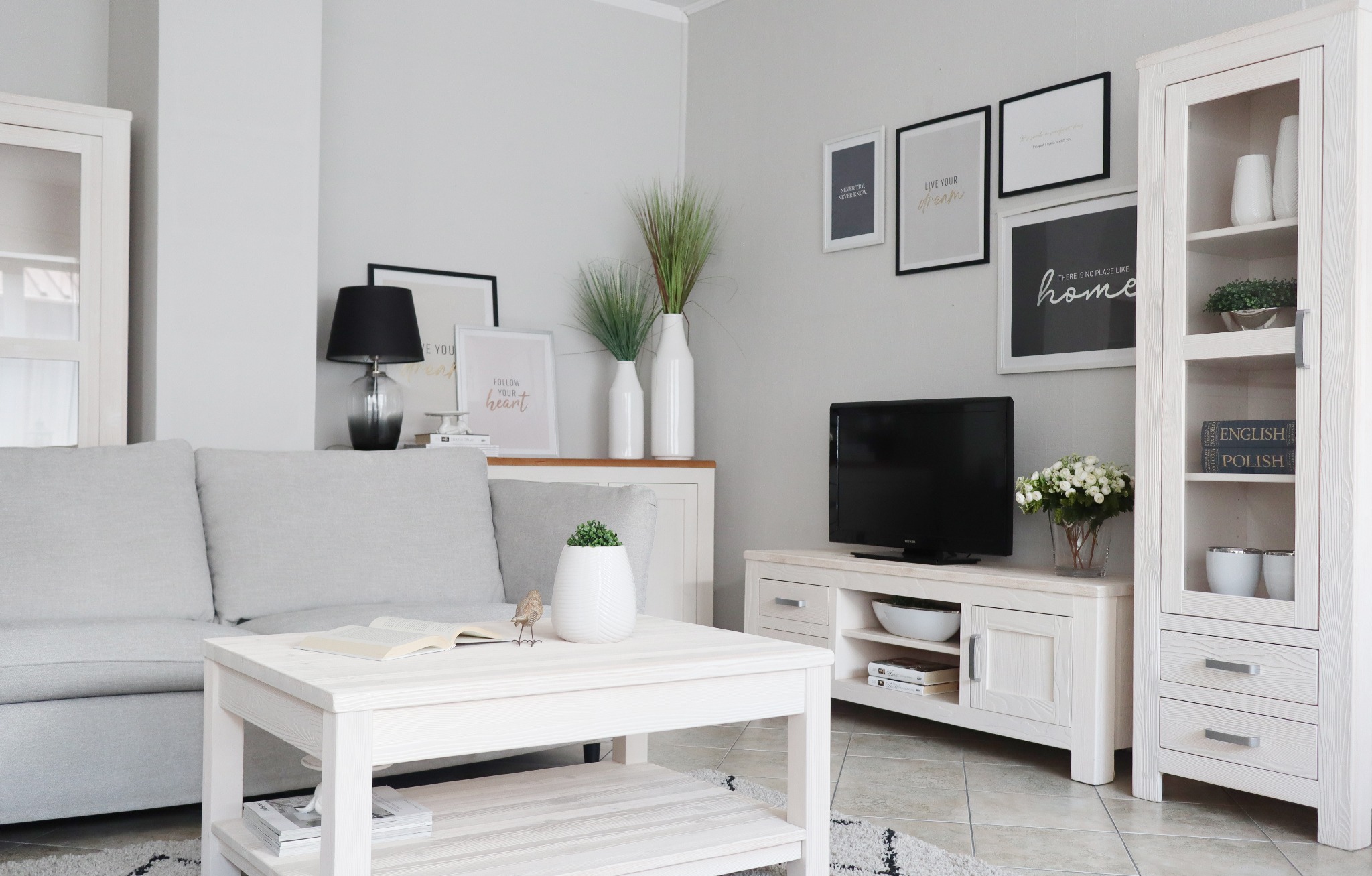 Jasne, przestronne wnętrze salonu z białymi meblami drewnianymi: szafka witrynowa, szafka RTV, stolik kawowy i sofa w szarym odcieniu. Na ścianie wiszą oprawione grafiki. Na stoliku leży otwarta...