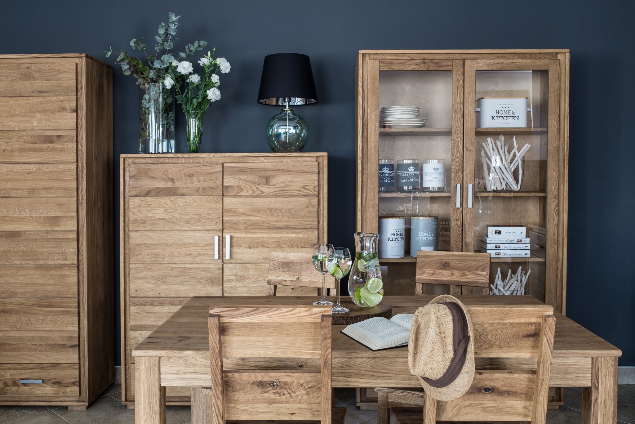 Komplet mebli drewnianych w jadalni: szafa, komoda z lampą, przeszklona witryna, stół z krzesłami, dzbanek z wodą i limonką, książka i kapelusz na krześle.