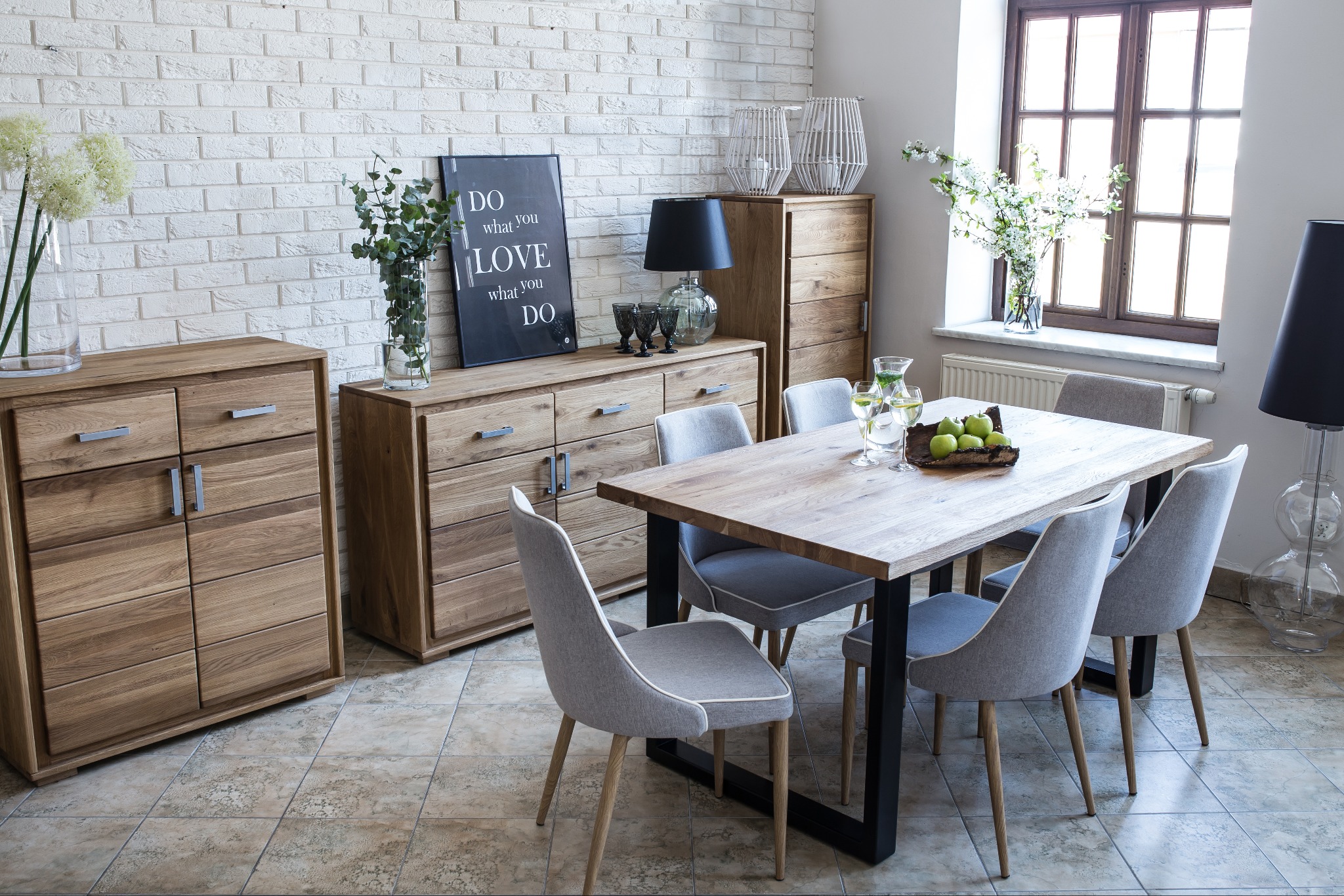 Jasne drewniane meble w jadalni z białej cegły: komoda z szufladami i szafka, stół z czarnymi nogami i szare krzesła, dekoracje i owoce na stole.