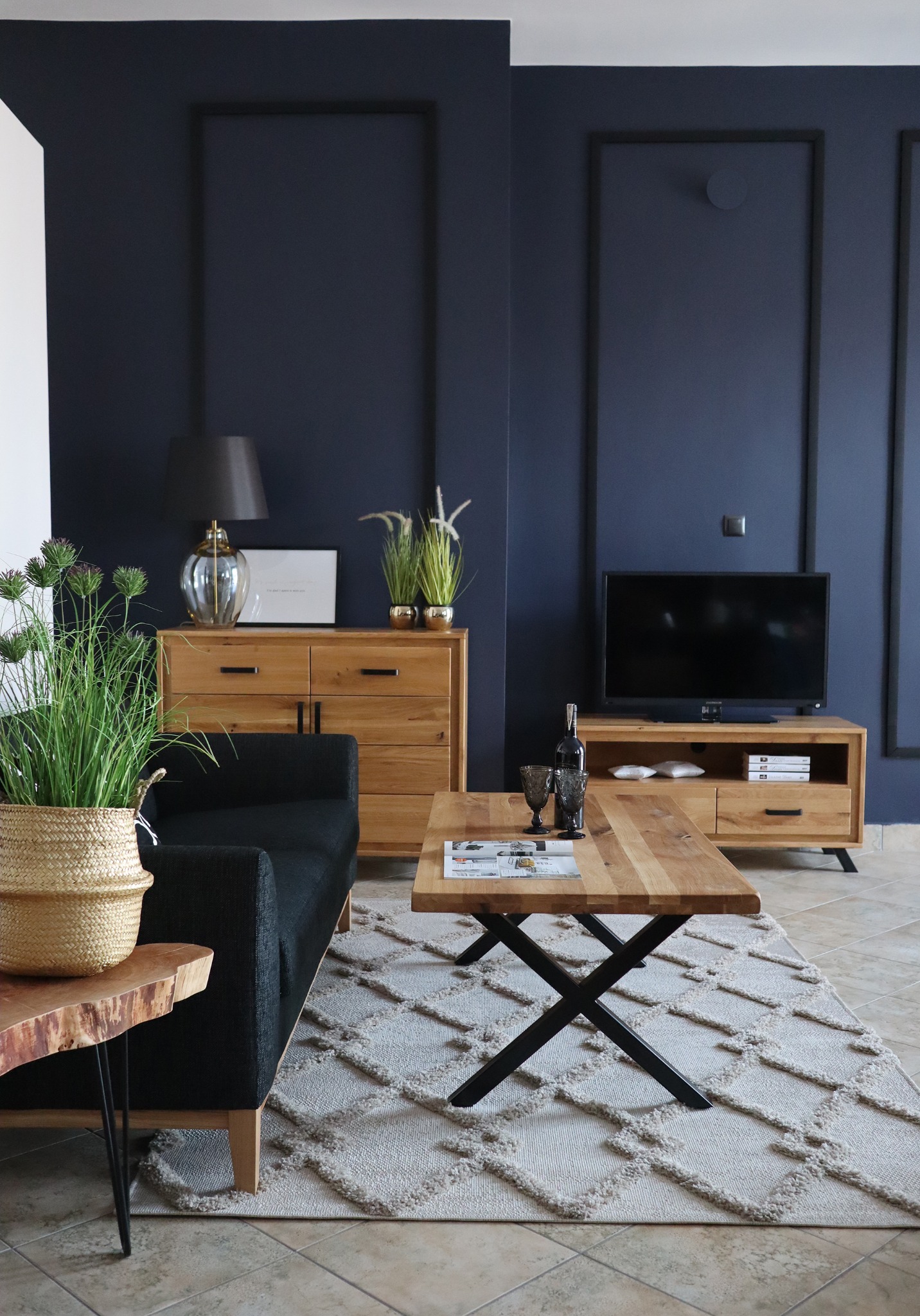 Elegancki salon z ciemnoniebieskimi ścianami w ozdobnych ramach, drewnianymi meblami, czarną sofą i dywanem w romby, oświetlony naturalnym światłem.