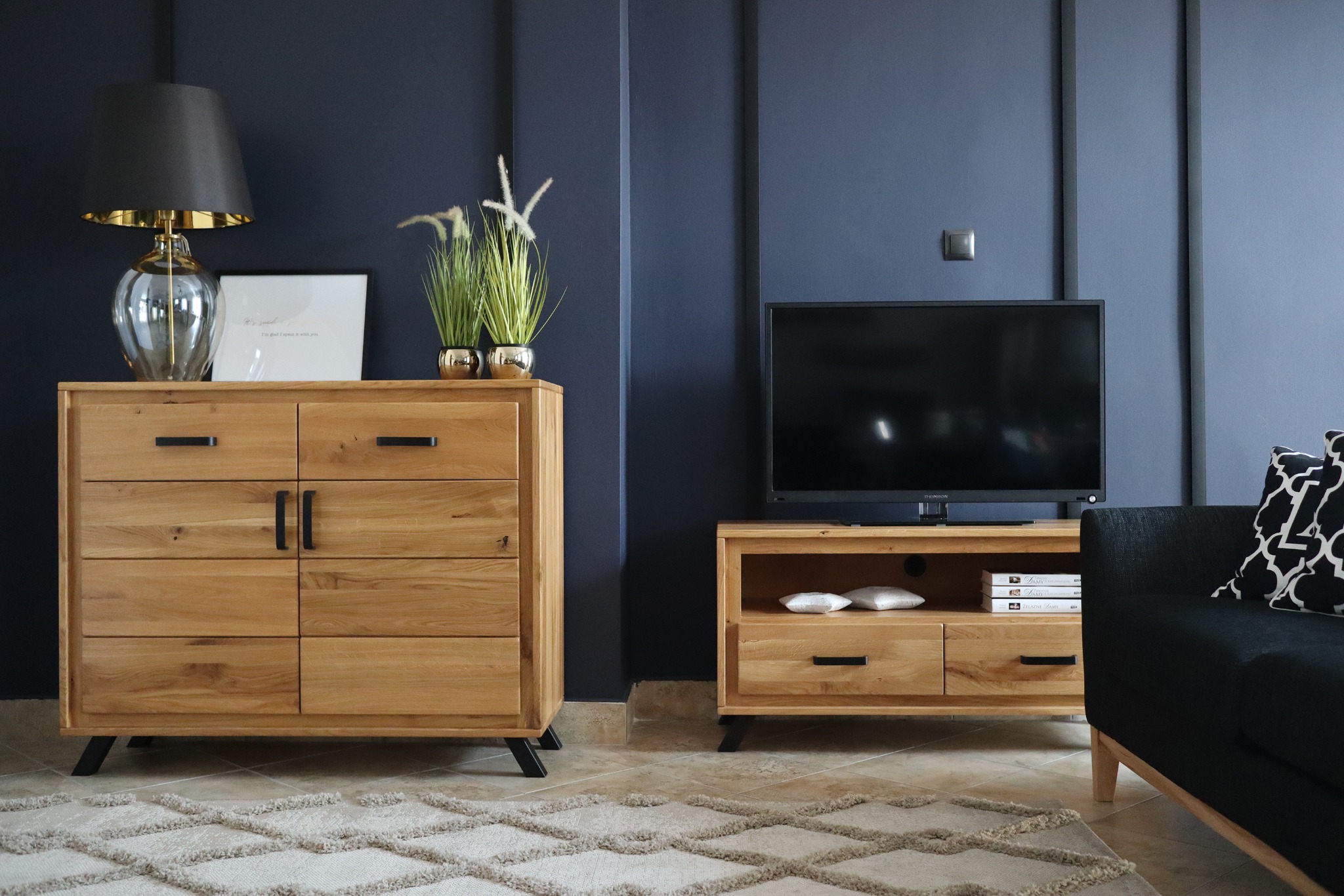 Drewniana komoda z sześcioma szufladami i czarnymi uchwytami, obok szafka RTV z telewizorem na tle granatowej ściany z dekoracyjnymi listwami, dywan w geometryczne wzory.