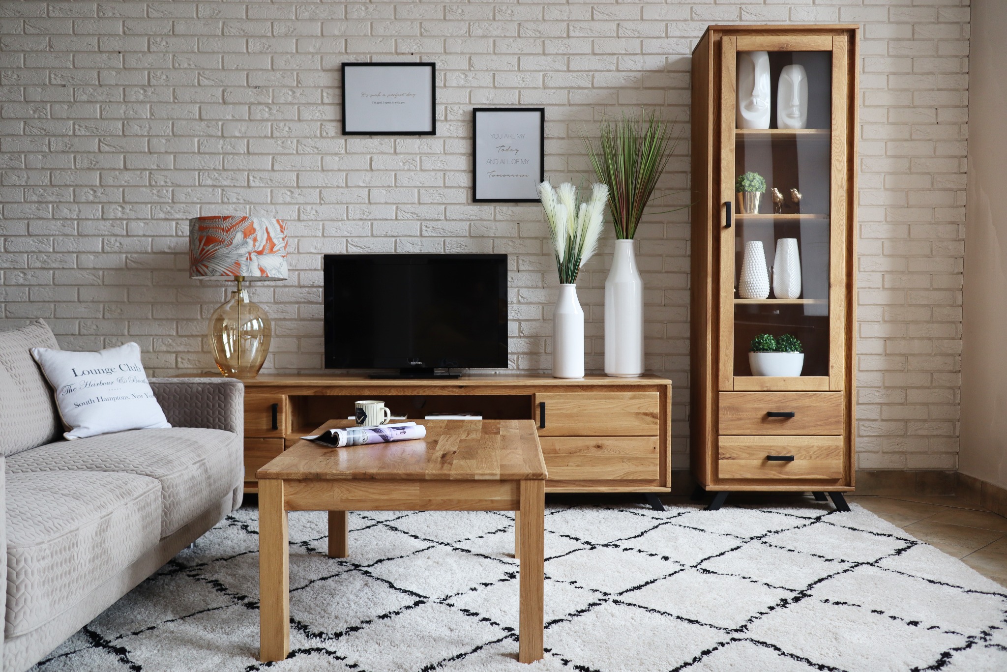 Jasny salon z drewnianymi meblami: szafka z przeszkleniem, szafka RTV i stolik kawowy na tle ściany z białej cegły i wzorzystego dywanu.