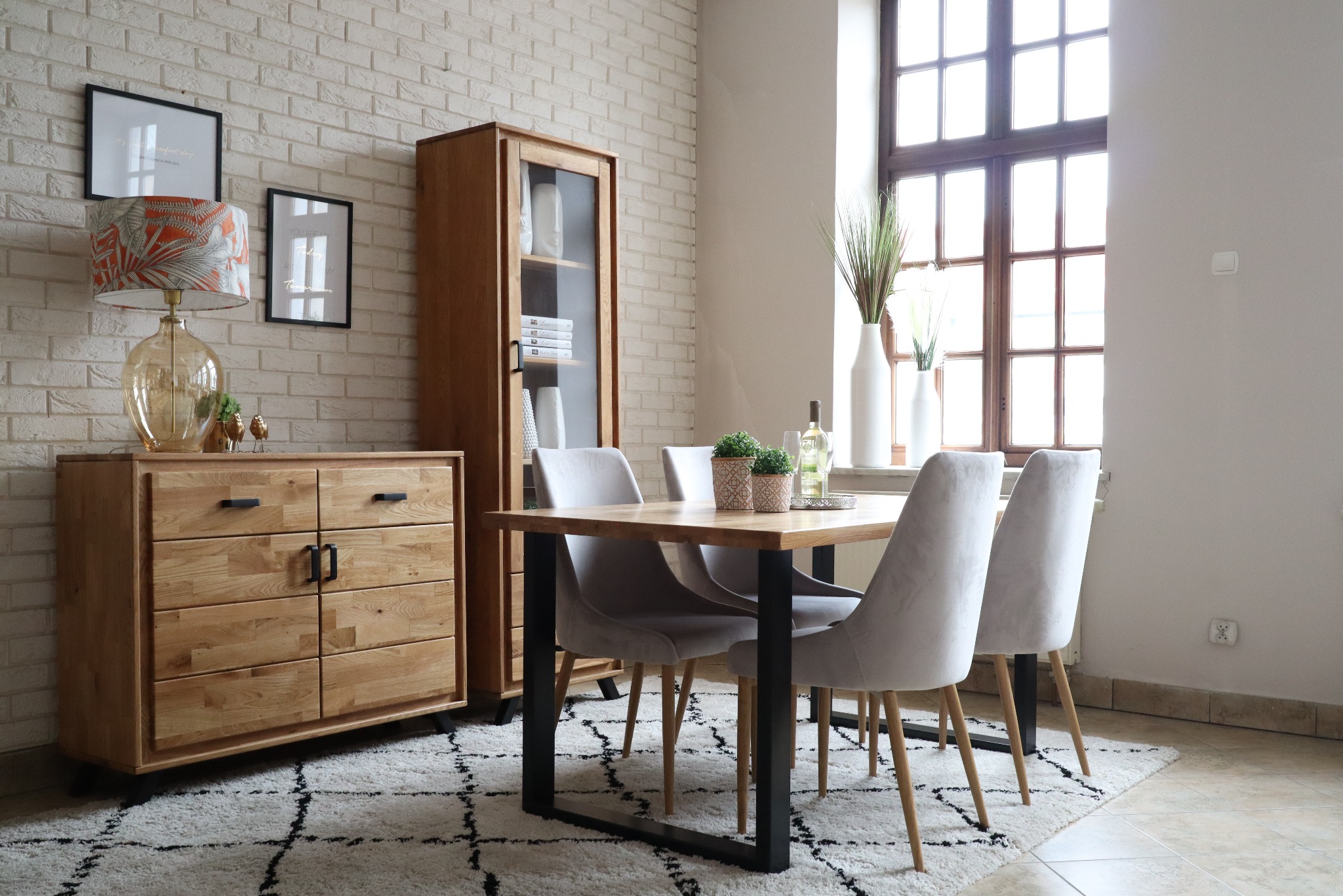 Jasne wnętrze jadalni z drewnianym stołem na czarnych nogach, obitymi szarymi krzesłami, komodą i przeszkloną witryną z jasnego drewna na tle ceglanej ściany i wzorzystego dywanu.