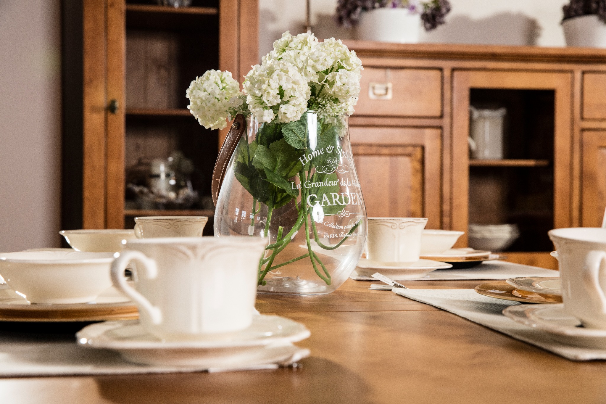 Ustawiony stół z porcelanową zastawą, wazonem z białymi kwiatami i drewnianą komodą w tle, tworzący ciepłą, domową atmosferę.