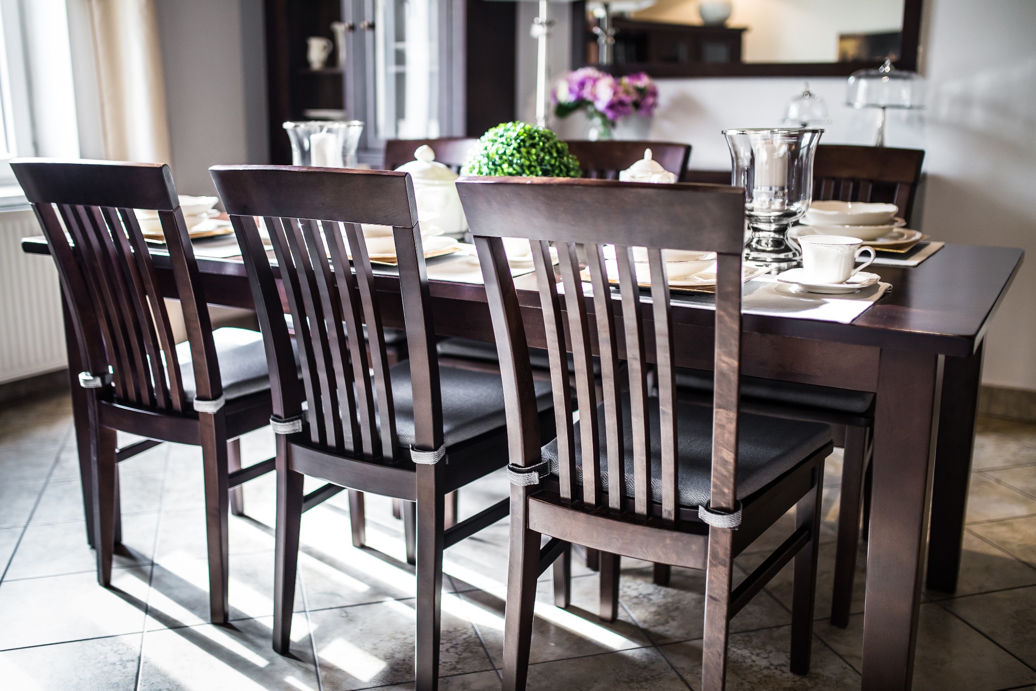 Ciemny, drewniany stół z nakryciem i krzesłami w jadalni, z dekoracyjnym bukietem kwiatów i elementami zastawy w tle.