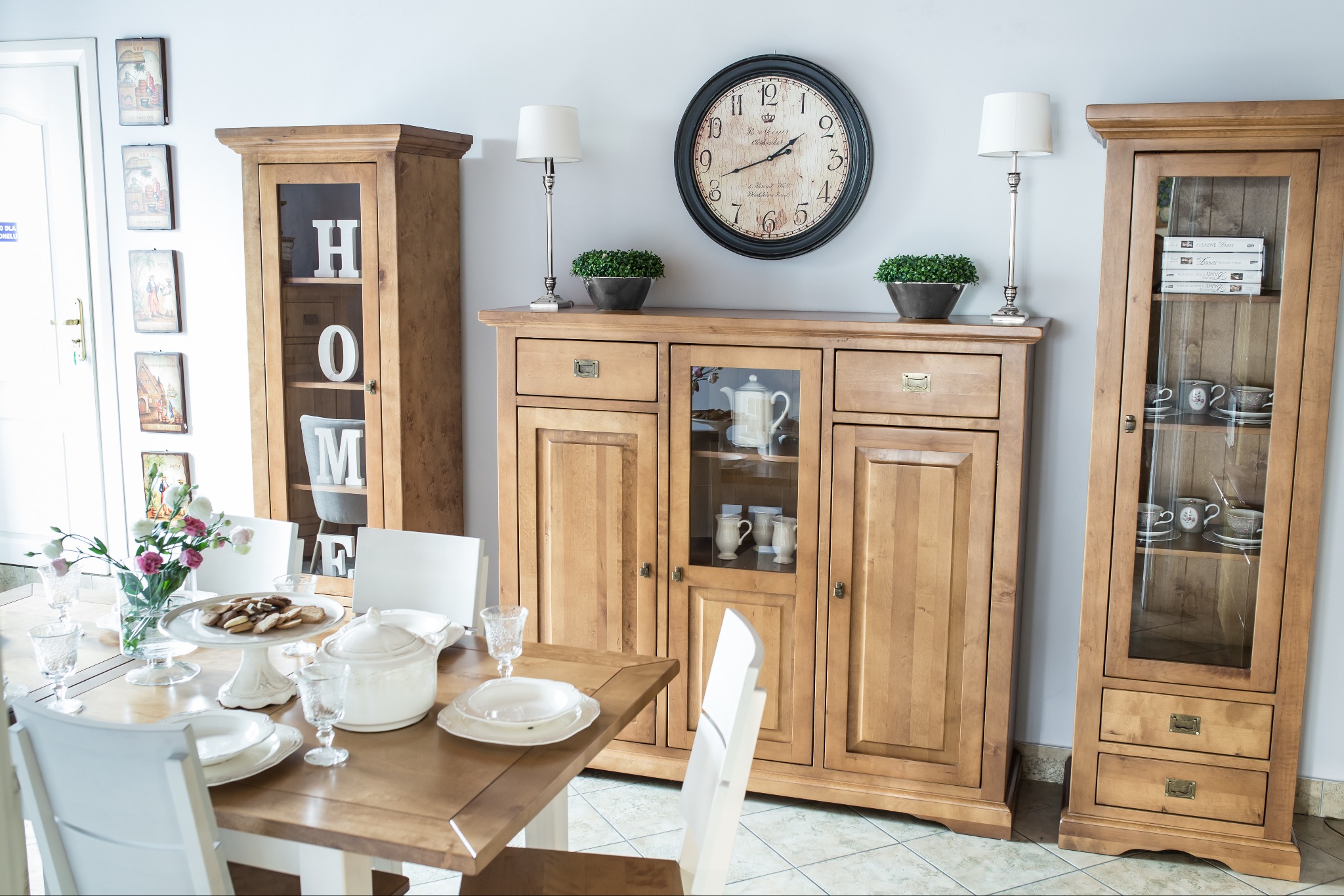 Drewniany kredens i witryny z przeszkleniami, uzupełnione dekoracyjnymi lampkami, zegarem i elementami zastawy stołowej w jadalni.