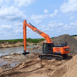 Schwer-Bud - Pomarańczowa koparka Develon na brzegu zbiornika wodnego, obok hałdy piasku, w tle zielone pole i błękitne niebo z chmurami.