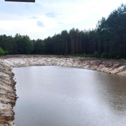 Schwer-Bud - Szeroki, świeżo wykopany staw z wodą, otoczony piaszczystym brzegiem. W tle zielony las i zabudowania. Widoczne ślady maszyn na brzegu stawu. Pochmurne niebo.