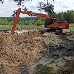Schwer-Bud - Pomarańczowa koparka Develon w trakcie prac ziemnych, widoczna świeżo przekopana ziemia, w tle las i drewniany budynek. Ujęcie spod gałęzi drzewa.