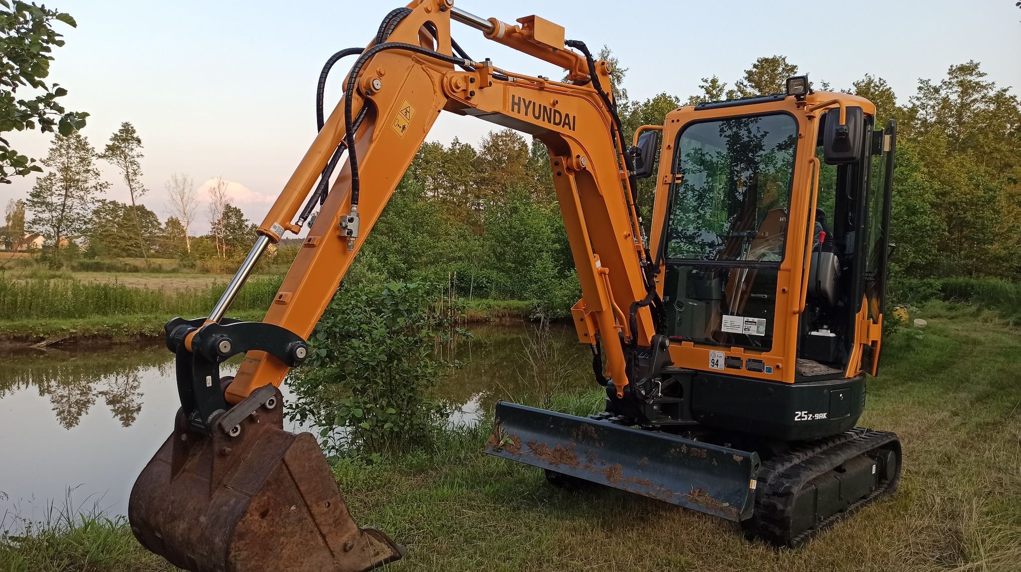 Żółto-czarna minikoparka Hyundai 25Z-9AK na trawiastym brzegu stawu, z widocznym łyżką i kabiną, na tle drzew i nieba o zmierzchu.