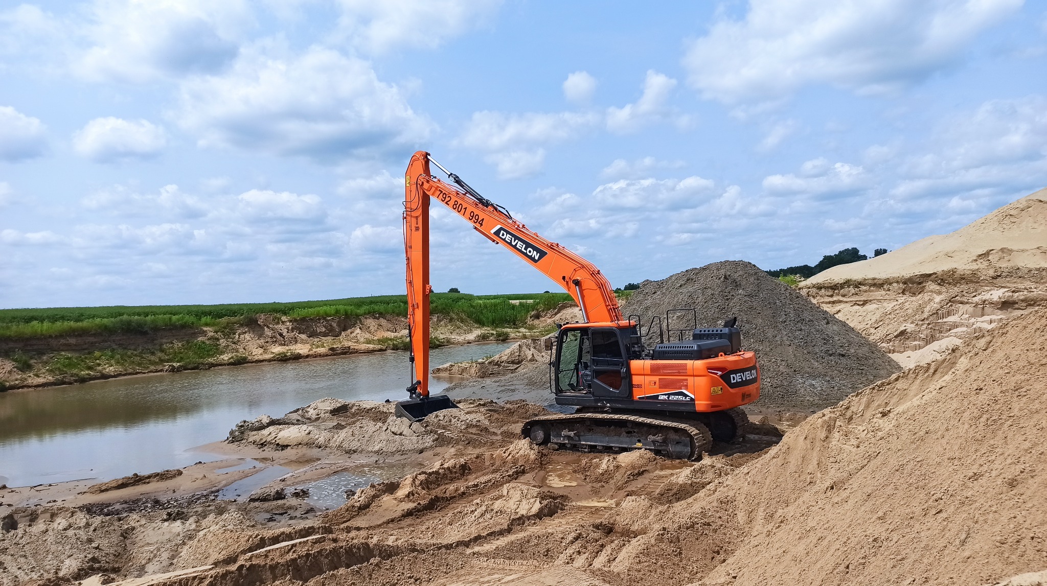 Pomarańczowa koparka Develon na brzegu zbiornika wodnego, obok hałdy piasku, w tle zielone pole i błękitne niebo z chmurami.
