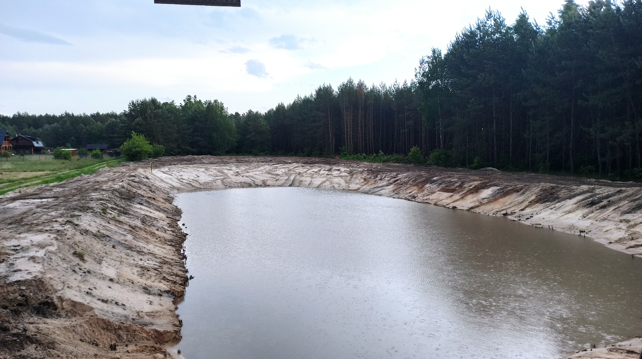 Szeroki, świeżo wykopany staw z wodą, otoczony piaszczystym brzegiem. W tle zielony las i zabudowania. Widoczne ślady maszyn na brzegu stawu. Pochmurne niebo.