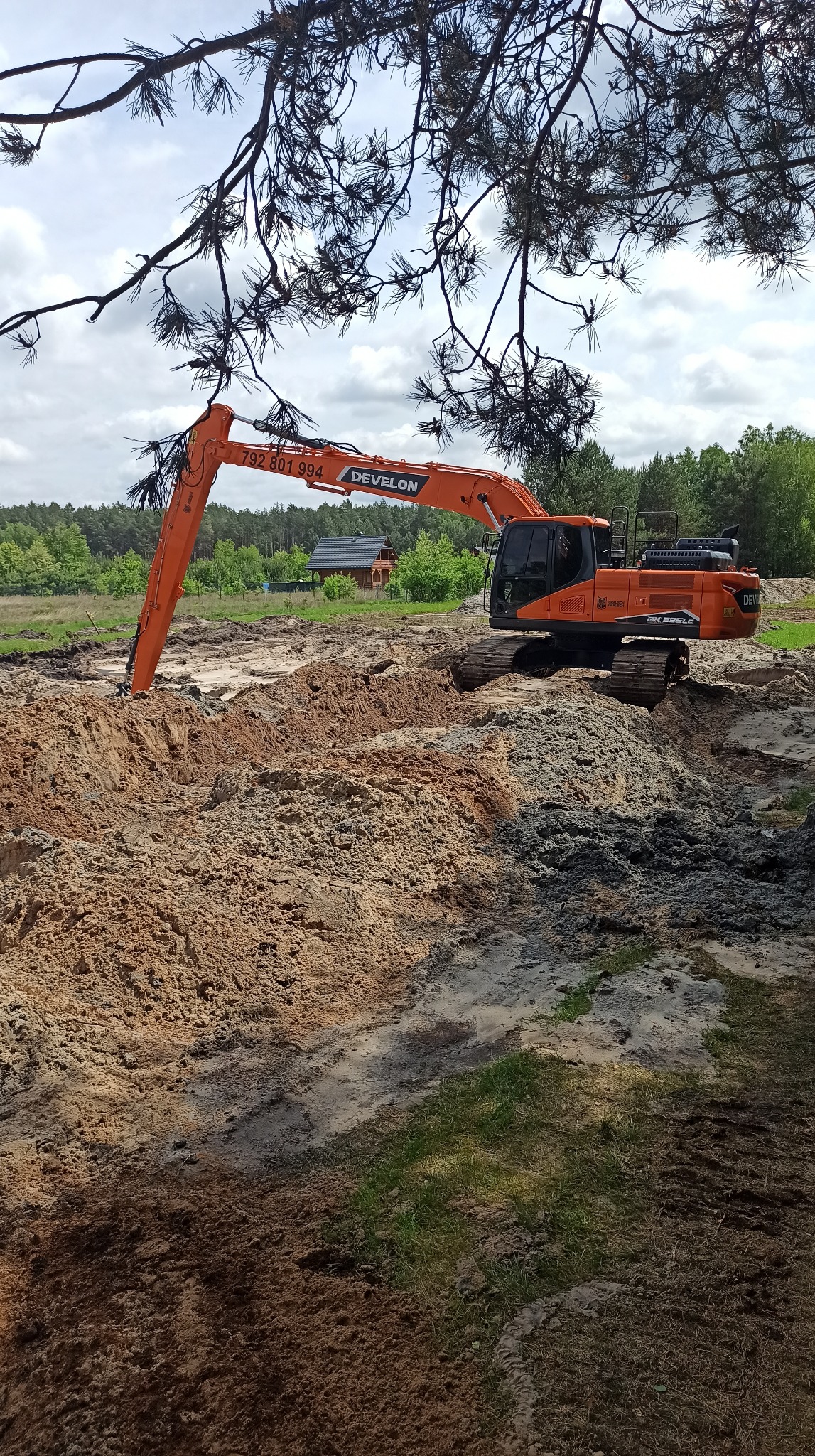 Pomarańczowa koparka Develon w trakcie prac ziemnych, widoczna świeżo przekopana ziemia, w tle las i drewniany budynek. Ujęcie spod gałęzi drzewa.