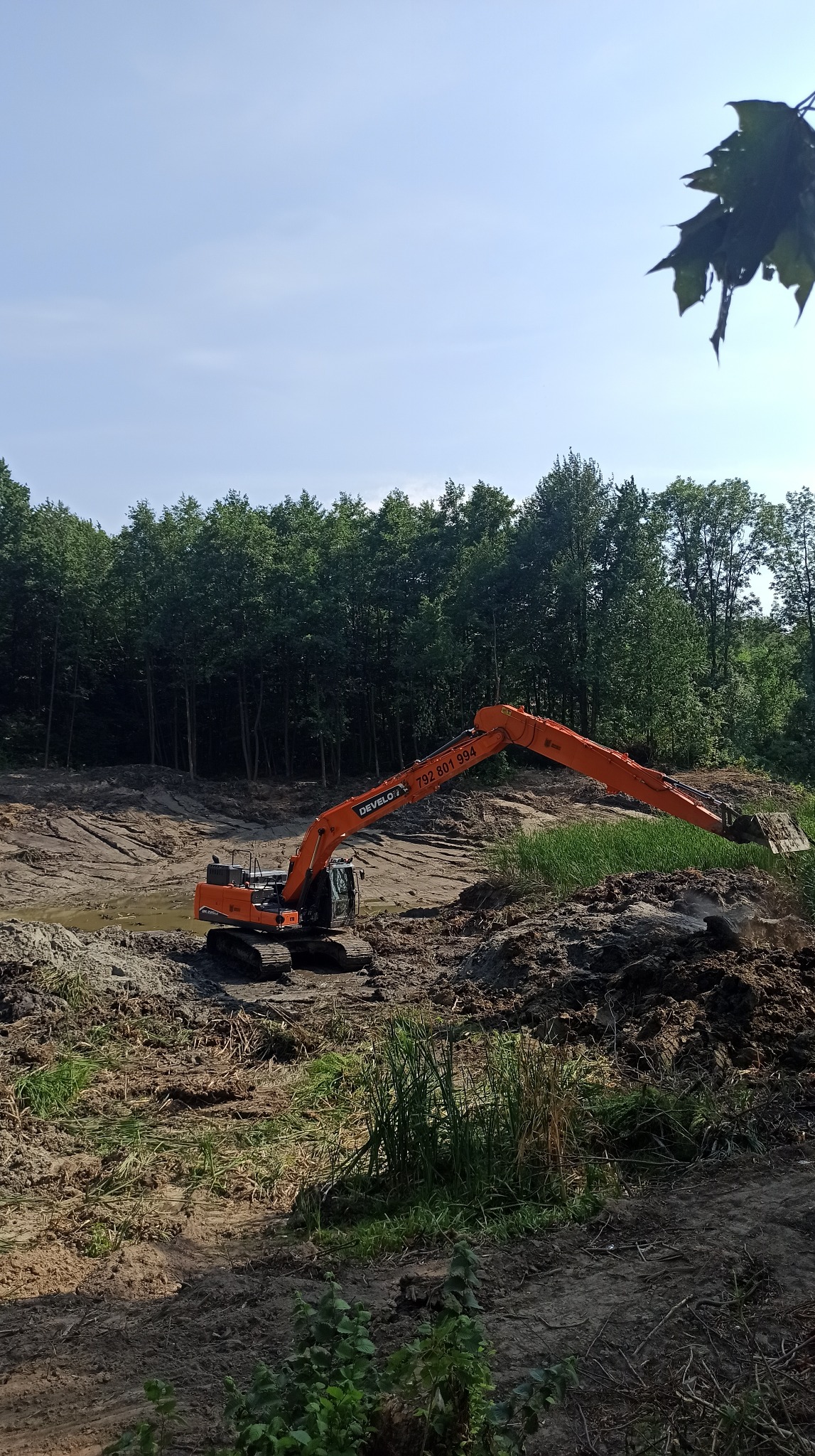 Pomarańczowa koparka w trakcie prac ziemnych na terenie z drzewami w tle. Widoczne świeżo przekopane błoto i wykopany dół.