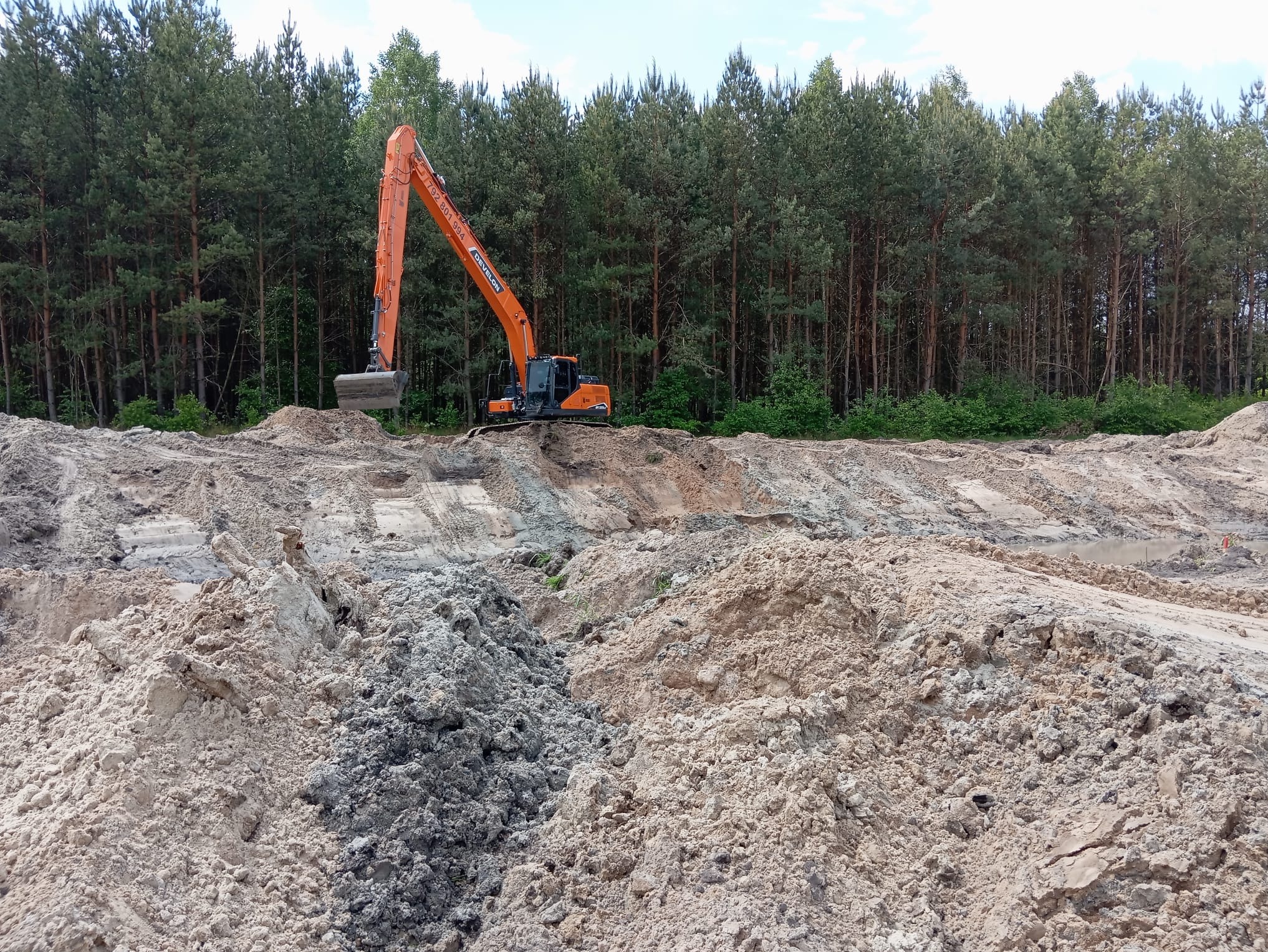 Pomarańczowa koparka Doosan na tle lasu podczas prac ziemnych, z widocznymi hałdami piasku i śladami opon. Widok na teren w trakcie wyrównywania.