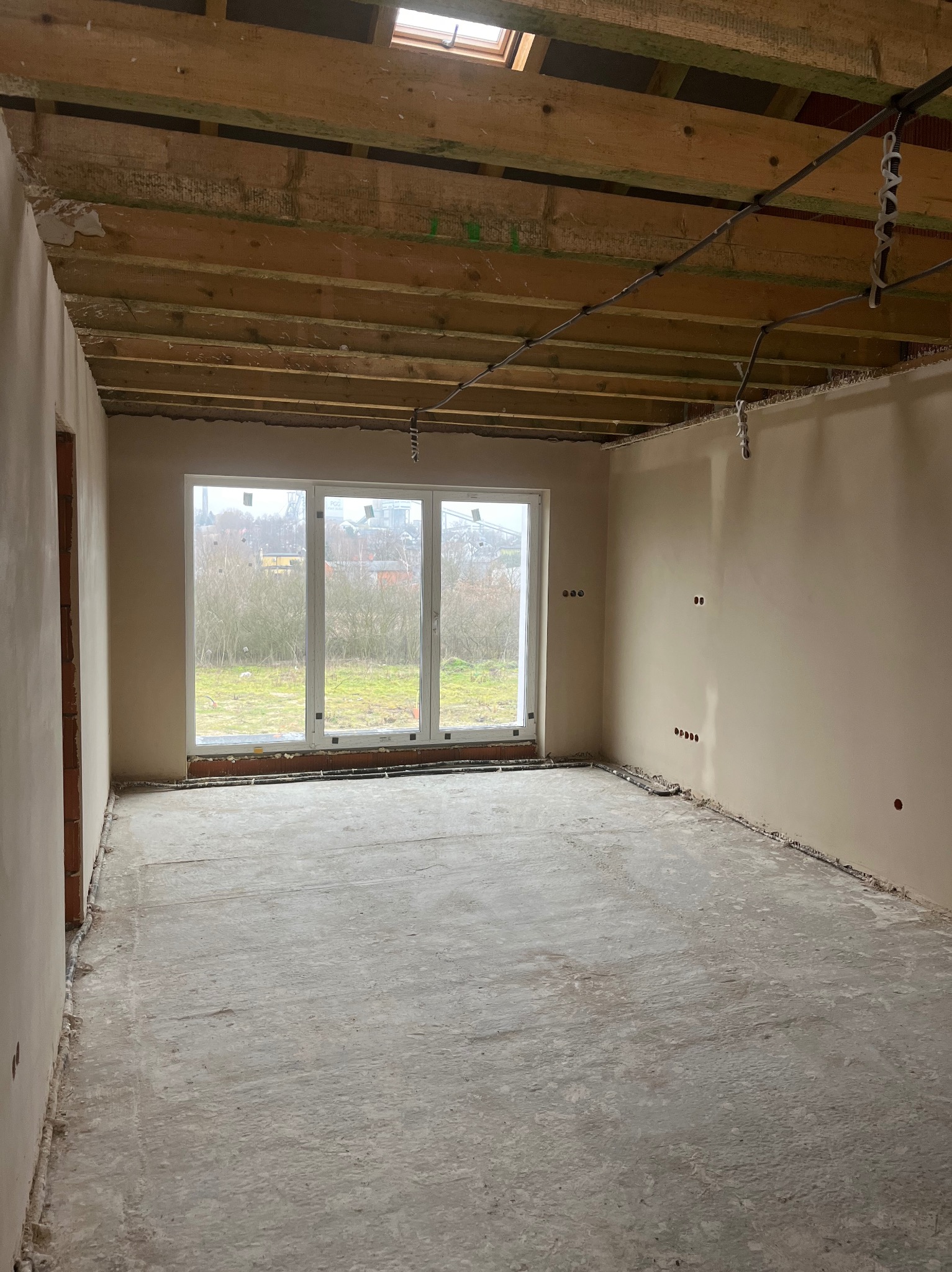 Wnętrze surowego pomieszczenia po nałożeniu tynków, widoczne otwory na gniazdka elektryczne, drewniana konstrukcja sufitu z oknem dachowym i okno z widokiem na zewnątrz.