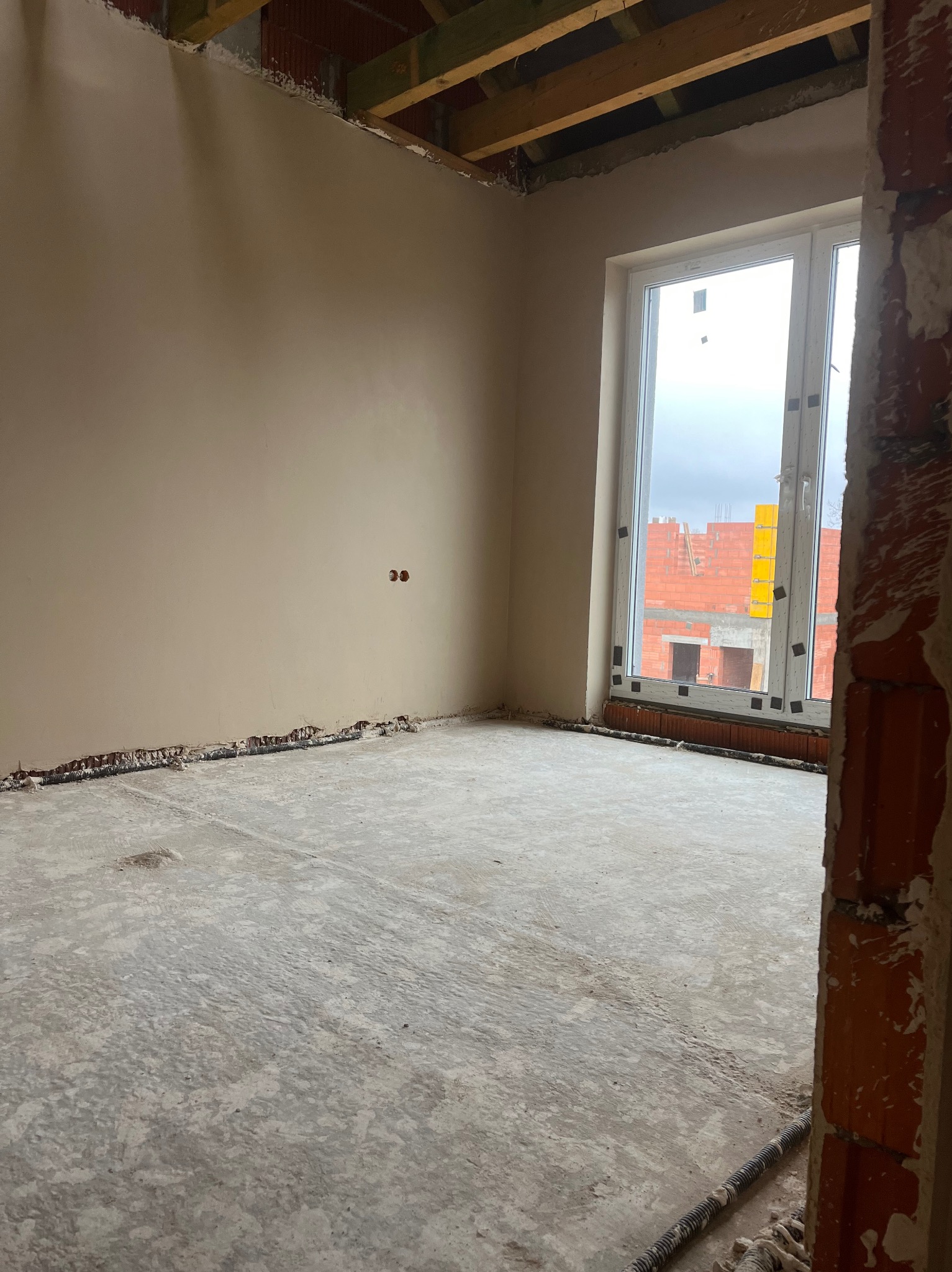 Wnętrze surowego pomieszczenia po nałożeniu tynku, widoczne ślady po instalacjach elektrycznych w ścianie, surowy beton na podłodze i niedokończone obramowanie okna z widokiem na budowę.