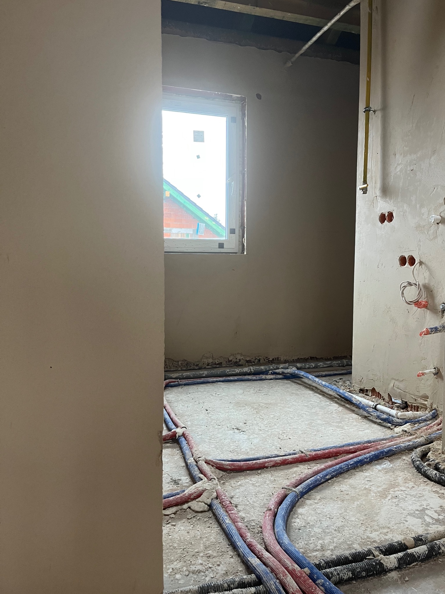 Świeżo otynkowane wnętrze z widocznymi przewodami instalacji wodnej i elektrycznej na podłodze przed wylewką, okno z widokiem na dach sąsiedniego budynku.
