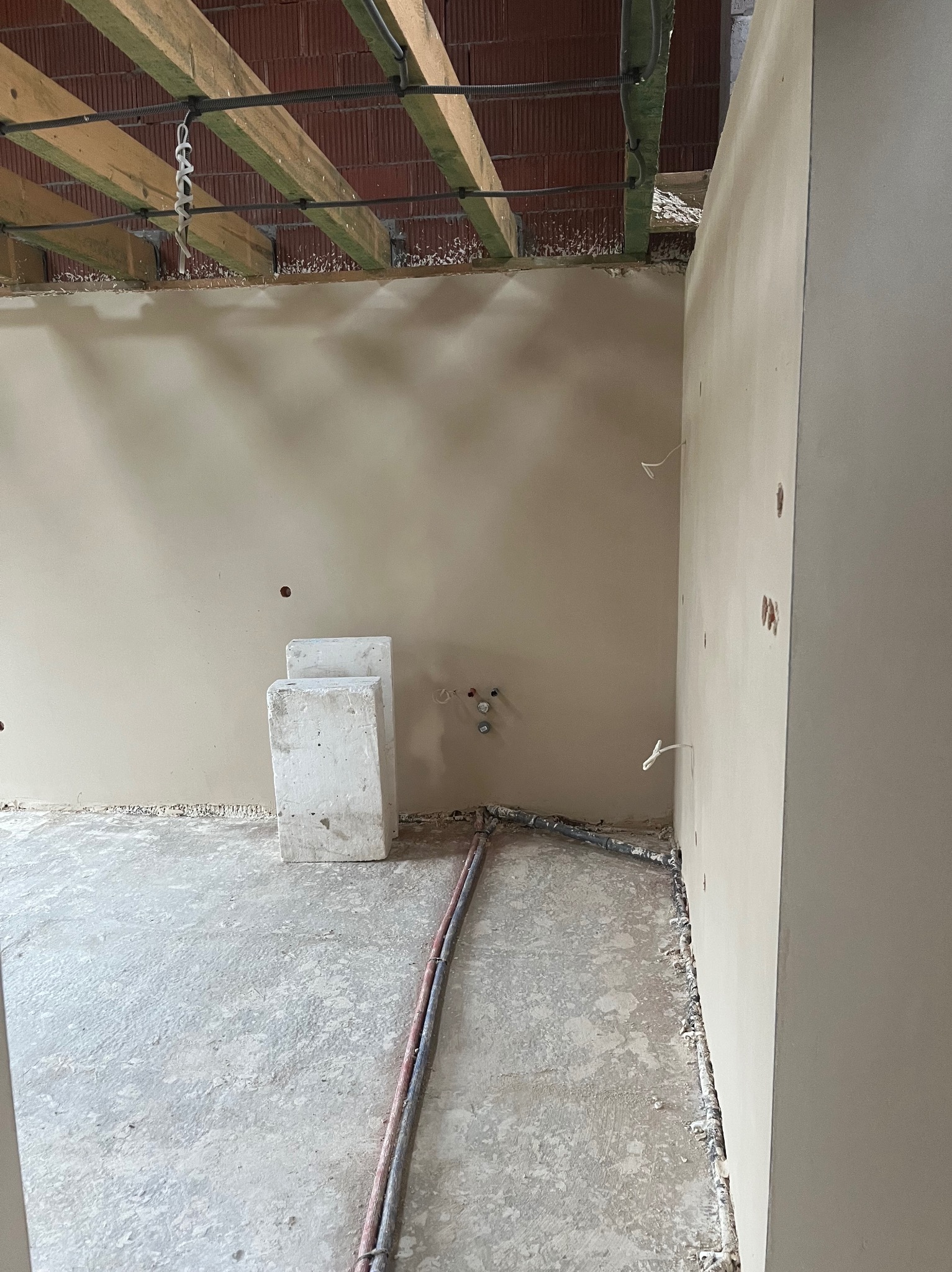 Wnętrze w trakcie prac wykończeniowych: gładkie, jasne ściany po nałożeniu tynku, widoczne wystające przewody instalacji elektrycznej oraz rury instalacji hydraulicznej przy podłodze, surowy...