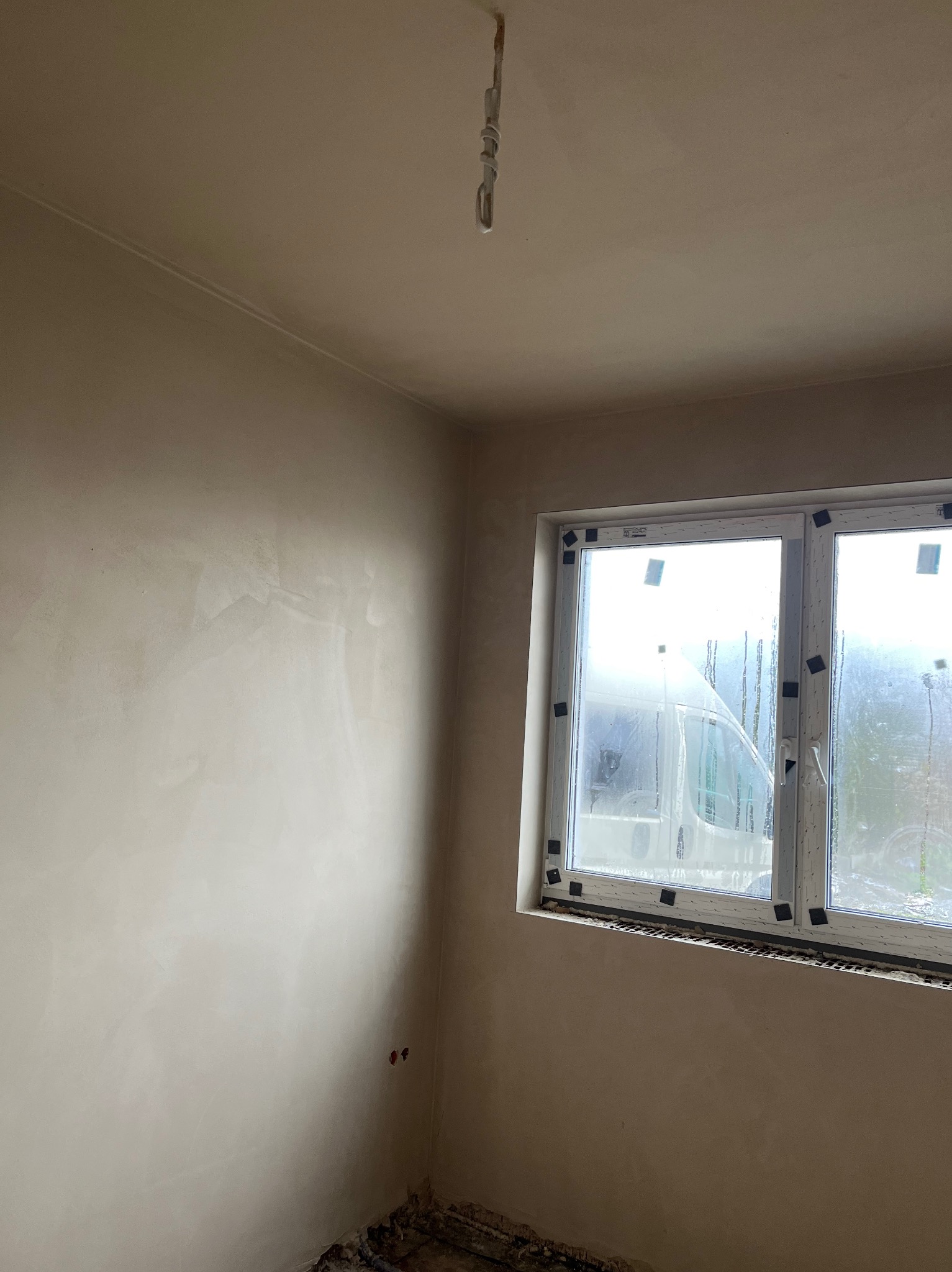 Świeżo otynkowane ściany w pokoju z oknem, widoczne ślady wilgoci na szybach i wystające kable z sufitu.