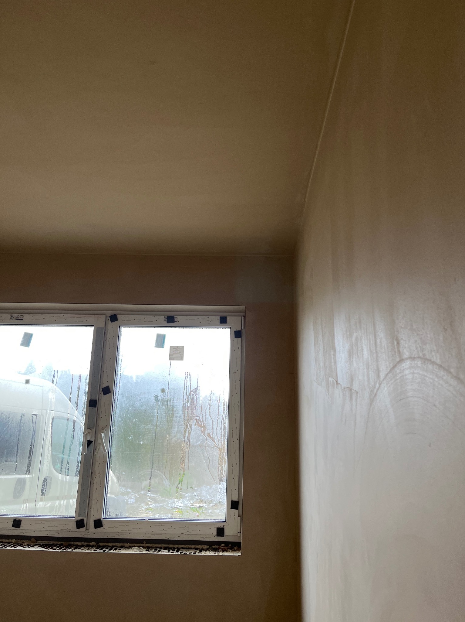 Świeżo nałożona, wilgotna warstwa gładzi na ścianie w rogu pomieszczenia, widoczne ślady po narzędziach, okno z zaparowanymi szybami i widocznym samochodem dostawczym na zewnątrz.