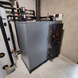 Mateusz Radomski - Szary piec na paliwo stałe marki Welding z czarnymi drzwiczkami, obok biały zbiornik akumulacyjny z czarnymi elementami, na tle beżowych płytek ściennych i podłogowych.