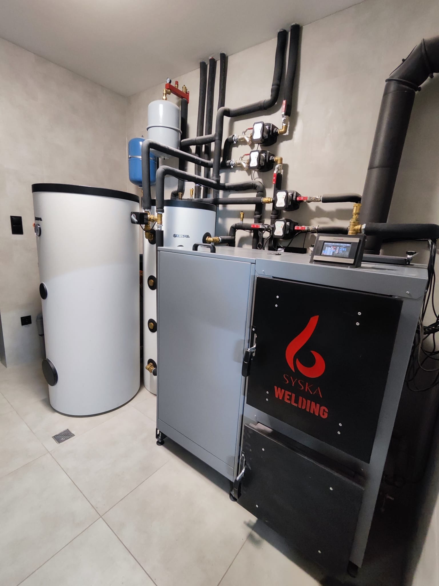 Nowoczesna kotłownia z piecem na paliwo stałe Syska Welding, zbiornikami buforowymi i instalacją hydrauliczną w minimalistycznym wnętrzu z szarymi płytkami.