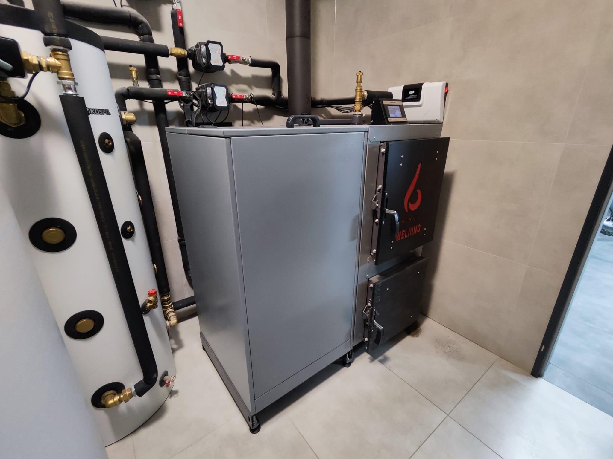 Szary piec na paliwo stałe marki Welding z czarnymi drzwiczkami, obok biały zbiornik akumulacyjny z czarnymi elementami, na tle beżowych płytek ściennych i podłogowych.