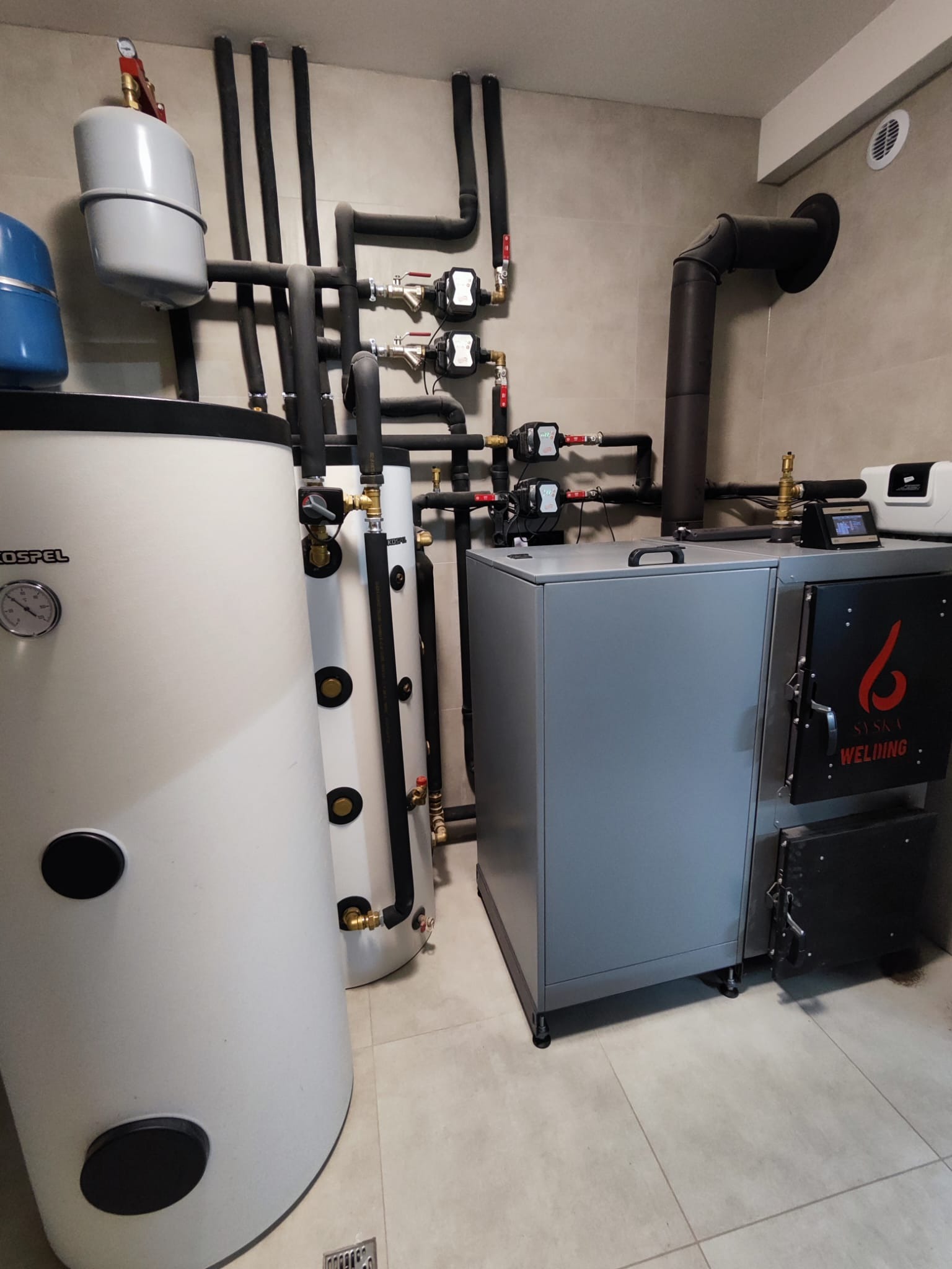 Szary piec na paliwo stałe marki Syska Welding z czarnym kominem, zbiorniki Kospel i instalacja CO w kotłowni wykończonej beżowymi płytkami.