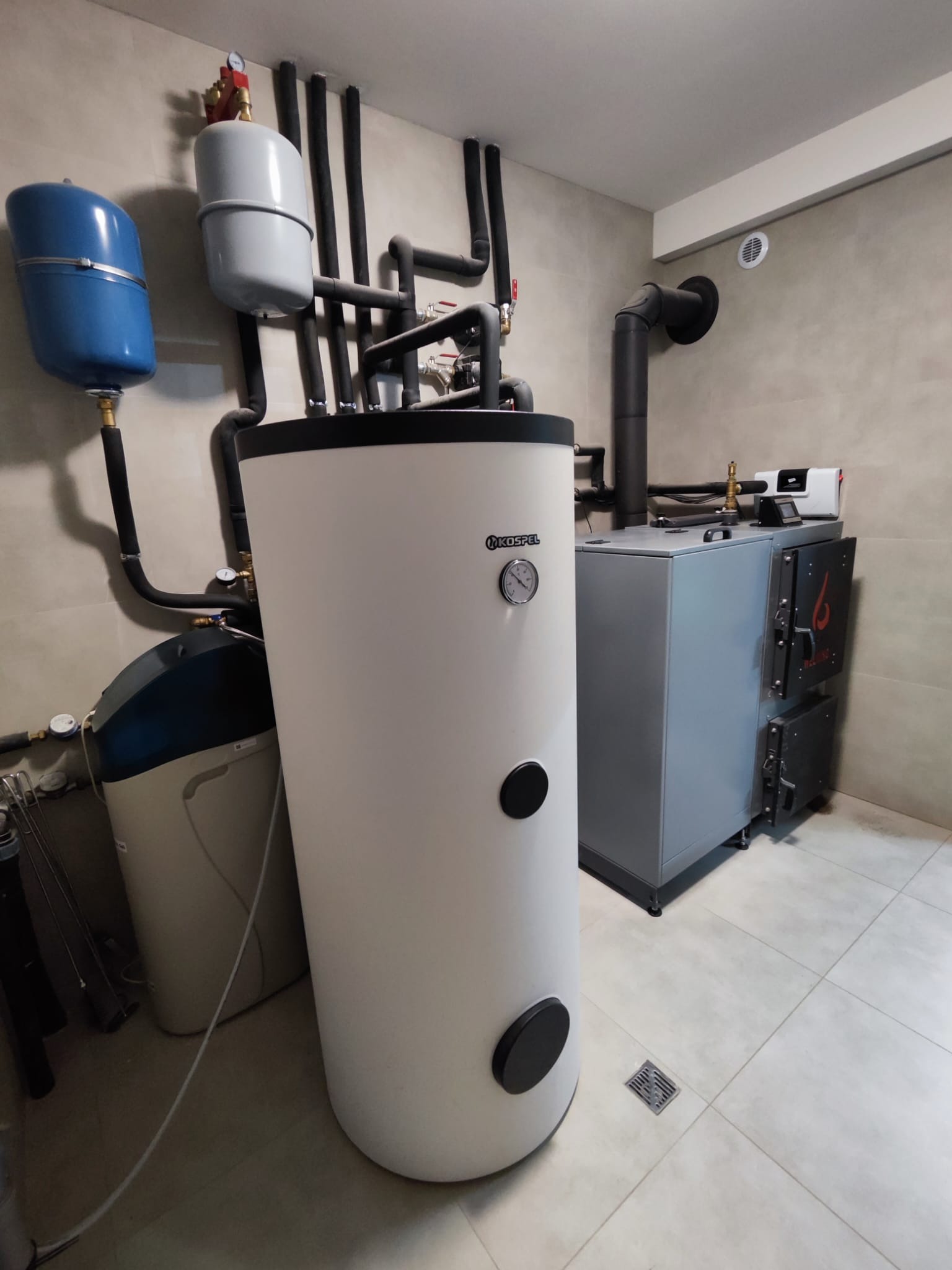 Biały piec Kospel z czarnymi elementami, obok szary piec z otwartymi drzwiczkami, rury instalacyjne na ścianie w jasnym pomieszczeniu.