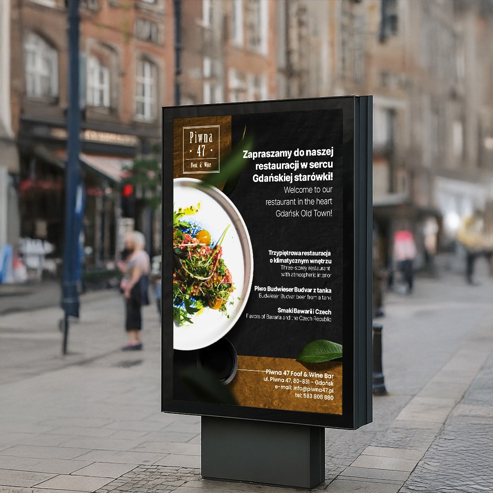 Billboard z reklamą restauracji Piwna 47 na tle ulicy w Gdańsku. Na plakacie danie i informacje o restauracji na Starym Mieście.