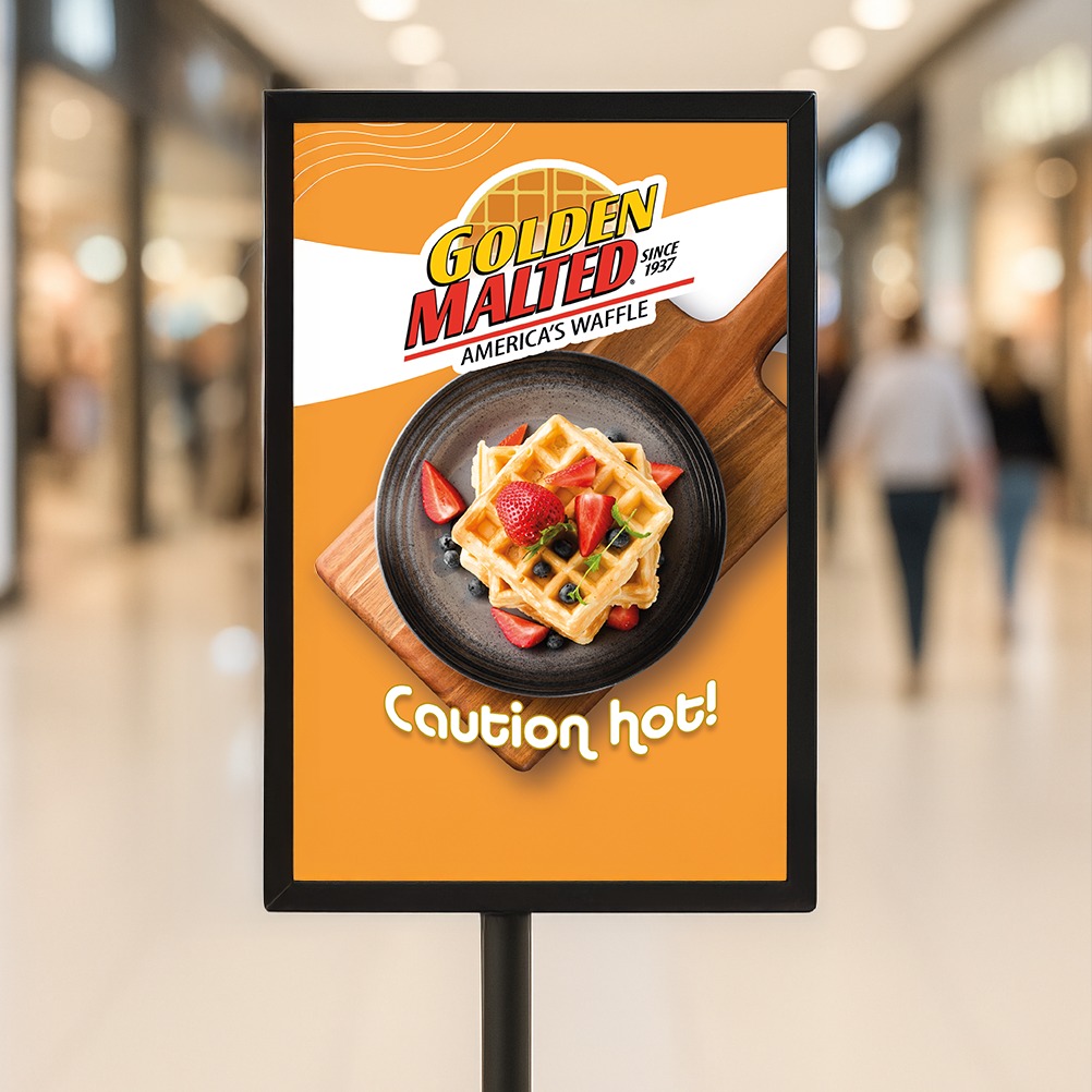 Stojak reklamowy z grafiką wafelków Golden Malted na tle rozmytego wnętrza centrum handlowego. Kolorystyka pomarańczowo-biała z ostrzeżeniem 'Caution hot!'.
