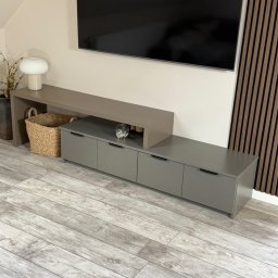 DOMAR - Szara szafka RTV na wymiar z szufladami i półką, dopasowana do wnętrza. Realizacja z Wrześni. Na blacie dekoracje, obok lamelki ścienne i kosz.