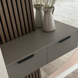 DOMAR - Szara szafka z dwiema szufladami i dekoracyjnym panelem z drewnianych listewek. Na blacie dwa wazony z suszonymi kwiatami. Minimalistyczny design.