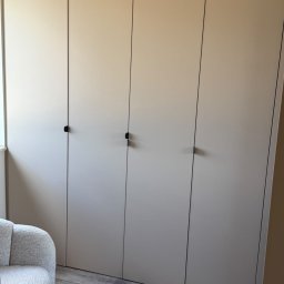 DOMAR - Beżowa szafa wnękowa z minimalistycznymi, czarnymi uchwytami, dopasowana do wnętrza z szarą podłogą i fotel w stylu bouclé. Nowoczesny design.