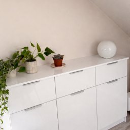 DOMAR - Biała, lakierowana komoda na wymiar we Wrześni, z sześcioma szufladami z minimalistycznymi uchwytami. Na blacie rośliny doniczkowe i okrągła lampa. Ustawiona na poddaszu.