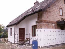 Ocieplanie fragmentu elewacji domu jednorodzinnego z widocznym styropianem i nieotynkowaną częścią budynku z cegły. Niebieska beczka i deski obok ściany.