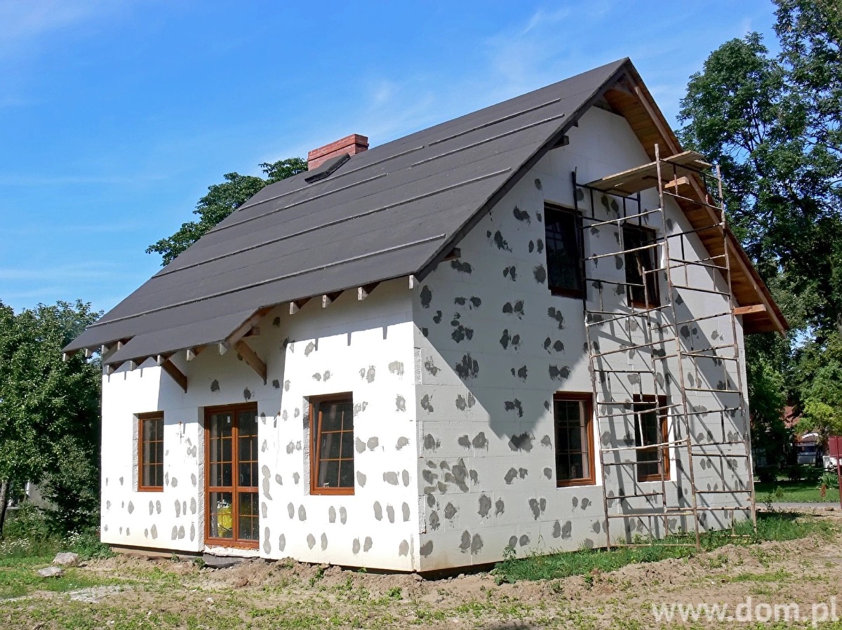 Dom w trakcie ocieplania, z widocznym styropianem i rusztowaniem przy oknie. Elewacja w stanie surowym, przygotowana do dalszych prac wykończeniowych.