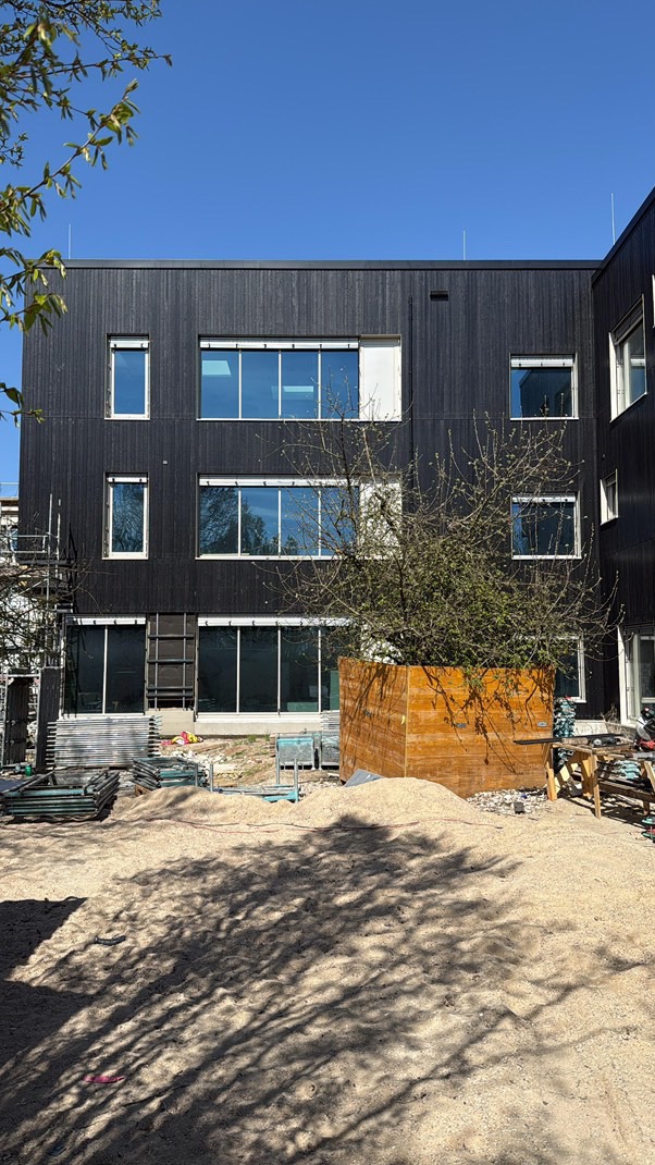 Nowoczesny budynek z ciemną, drewnianą elewacją i jasnymi oknami. Widoczne rusztowania i materiały budowlane na placu przed budynkiem.