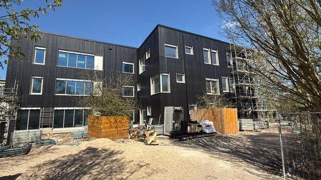 Nowoczesny budynek z czarną drewnianą elewacją i białymi oknami, częściowo w rusztowaniach. Widoczne materiały budowlane i przygotowany teren wokół budynku.